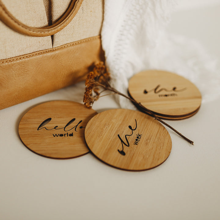 A round dark bamboo set of milestone cards on a white blanket. The three visible ones say hello world, one week and one month.