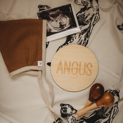 Light bamboo 'Borderline Plaque' in 14cm size, surrounded by baby paraphernalia: a scan photo, wooden rattles and a bonnet on bear-printed blanket.