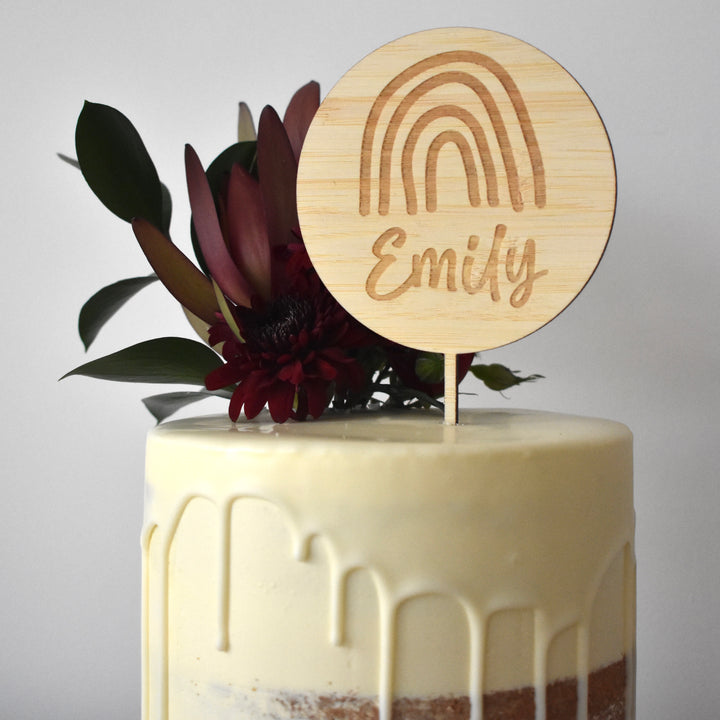 A white drip cake with dark red flowers and greenery is topped with the Lovely Lettering Rainbow Cake Topper (other colours available), featuring a hand-drawn rainbow and the name Emily for a charming touch.