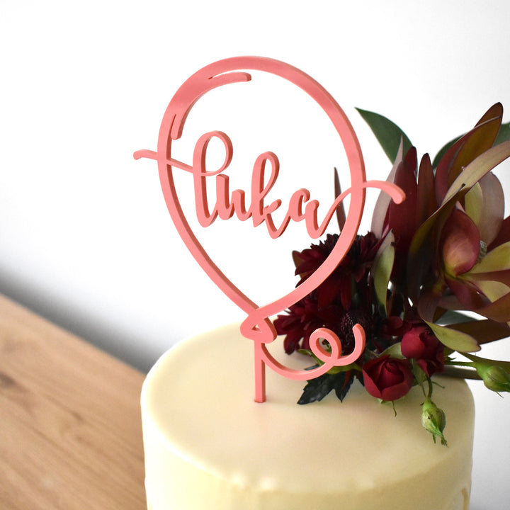A pink acrylic balloon shaped cake topper, with the name Luka in linked writing inside. Sitting atop a white cake with floral arrangement.