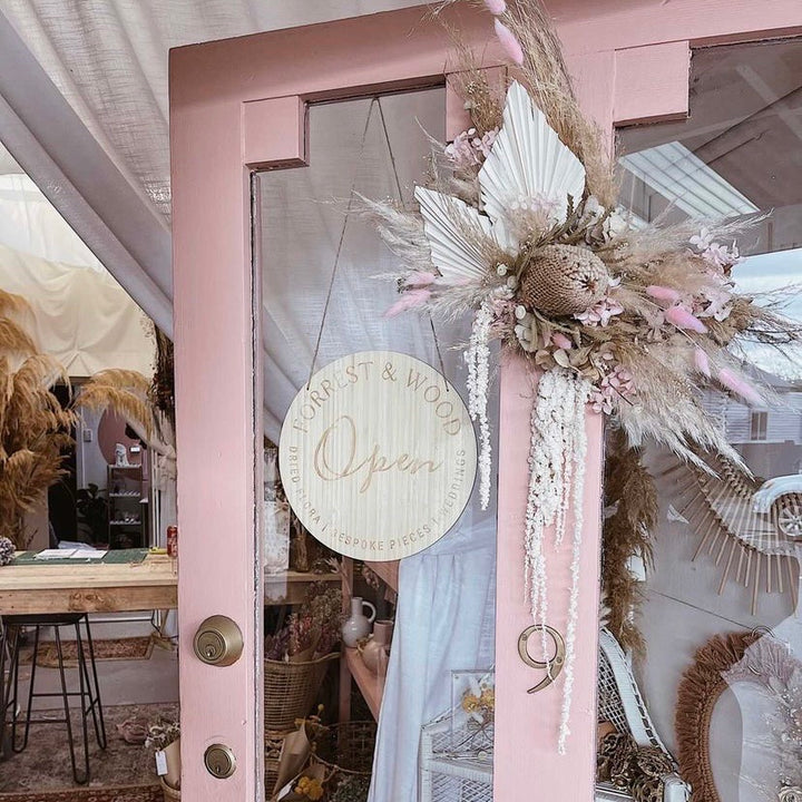 A light bamboo round sign saying open, with Forrest and Wood logo engraved. Hanging on a pink door with a dried floral arrangement.