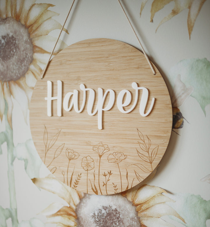 The Personalised Botanical Layered Plaque features “Harper” in white script with engraved flowers on a round wooden sign, hung by string—ideal as custom home décor, shown against a wall with a painted sunflower design.