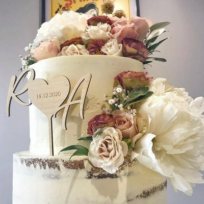 A light bamboo cake topper in the shape of heart with the letters R and A on either side. Etched inside the heart is 19.12.2020. Sitting on a white two layer cake with floral arrangement. 