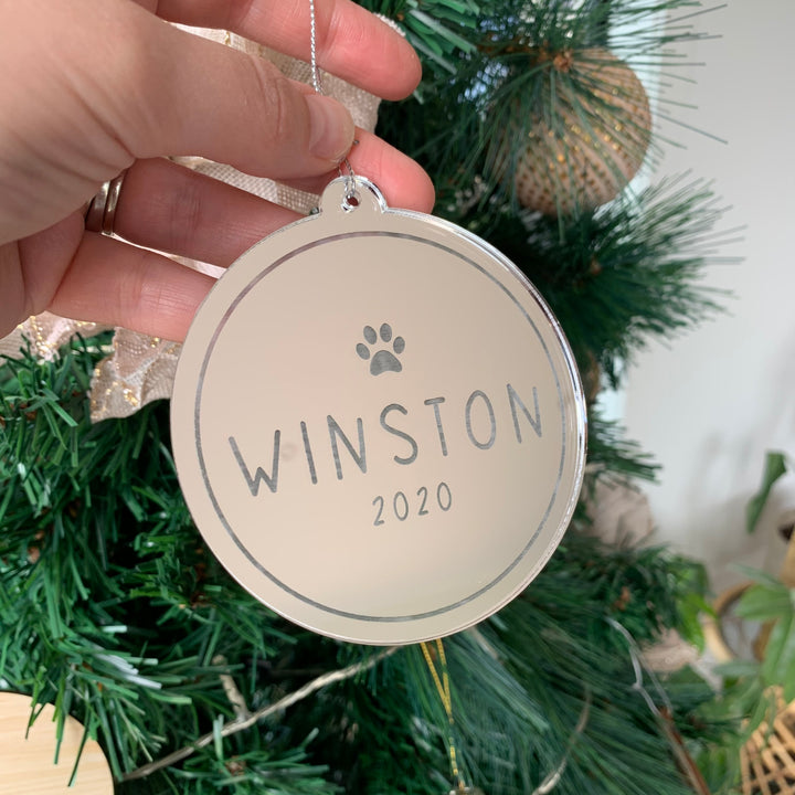 A hand holds an Etched Pets Christmas Bauble, a round, reflective ornament with a paw print and personalised text, displayed in front of a decorated green Christmas tree.