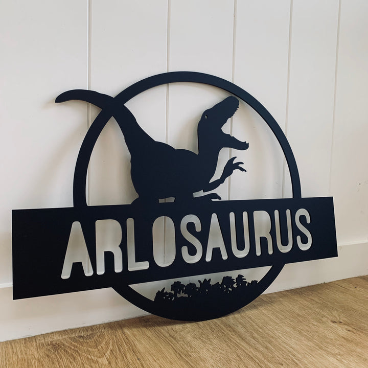 A Personalised Dinosaur Wall Plaque, crafted from black metal and shaped like a circular logo with a dinosaur silhouette and cut-out letters, leans against a white paneled wall on a wooden floor.
