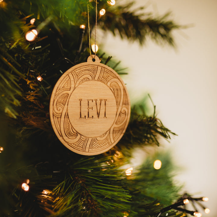 A close-up of a wooden Whānau Kirihimete Bauble engraved with LEVI, hanging on a green tree amid string lights and blurred branches—a unique ornament from Ani Huia Art&