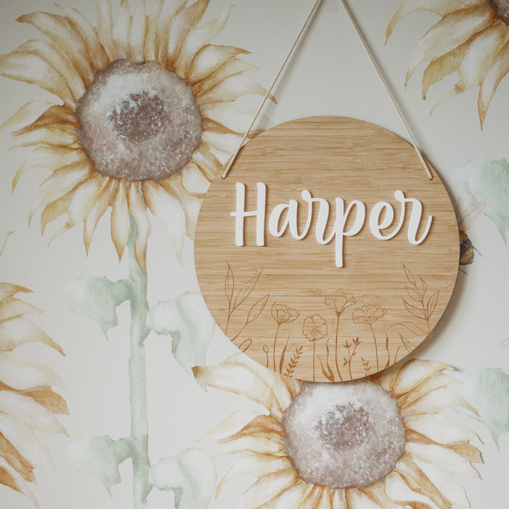 A Personalised Botanical Layered Plaque reading Harper hangs on a wall adorned with painted sunflowers and green stems, featuring a subtle floral engraving along the bottom edge for added charm.