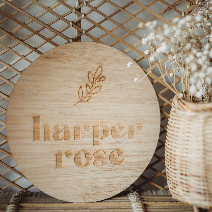The Personalised La Petite Stem Plaque, a round bamboo sign with an engraved branch and custom text, rests against a woven lattice near a wicker basket of dried white flowers—a charming birth announcement or personalised display.