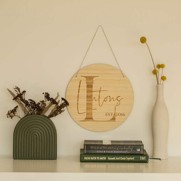 A Personalised Monogram Plaque reading The Hintons EST 2016 hangs above a shelf with a green arch vase of dried flowers, a white vase with yellow billy balls, and three stacked books.