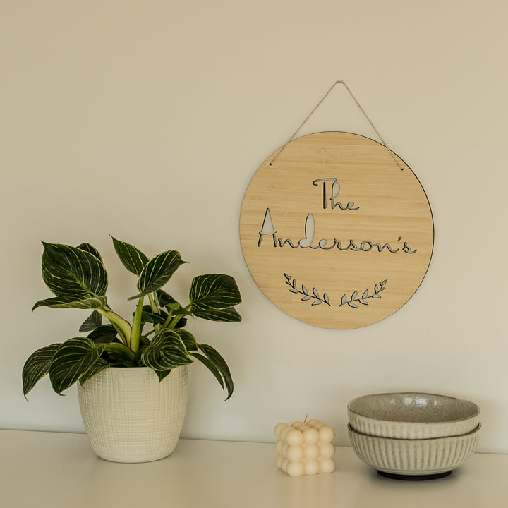 A Personalised Surname Plaque reading The Andersons hangs on a beige wall above a white surface with a potted plant, decorative candle, and stacked grey bowls.