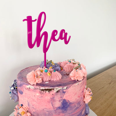 A pink and purple watercolour cake with sprinkles and a pink acrylic cake topper saying Thea.