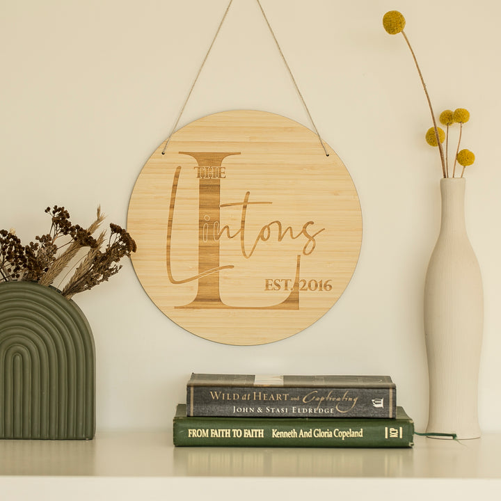 A round Personalised Monogram Plaque with "The Lintons EST. 2016" hangs above a shelf with dried flowers, a white vase holding yellow billy balls, and three stacked books—an ideal engagement gift or custom bamboo home decor piece.