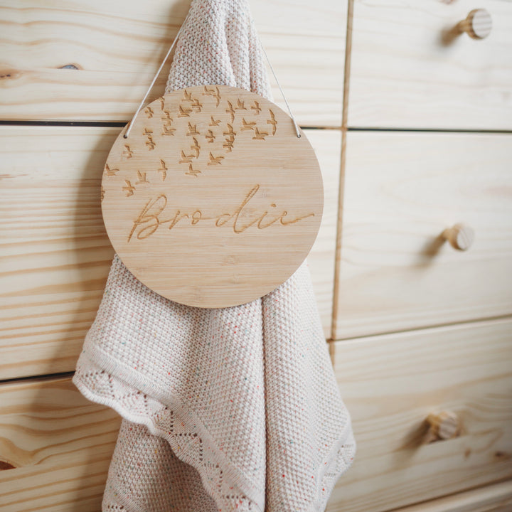 A round bamboo plaque with a flock of birds and the name Brodie etched on. Hanging from a chest of drawers.