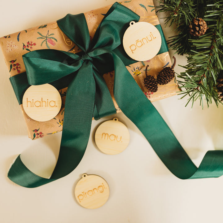A gift wrapped in patterned paper with a dark green ribbon is displayed with four round bamboo "Te Reo - Etched Want, Need, Wear, Read Tags (Unpersonalised)" beside pine sprigs and pinecones.