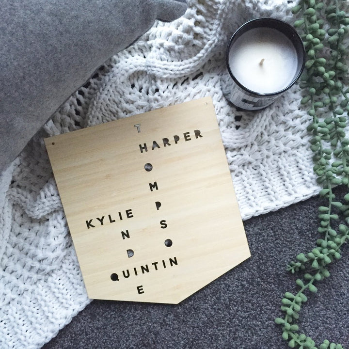 The Personalised Family Ladder, a bamboo board with names HARPER, THOMPSON, KYLIE, and QUINTIN in a crossword style, rests on a white knit blanket near a candle and green trailing plant for unique wall art decor.