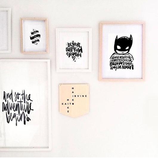 A white wall features the “Personalised Family Ladder” art piece, framed in a simple light-colored frame, alongside other art with handwritten text, abstract shapes, quotes, a crossword design, and an illustrated superhero mask.