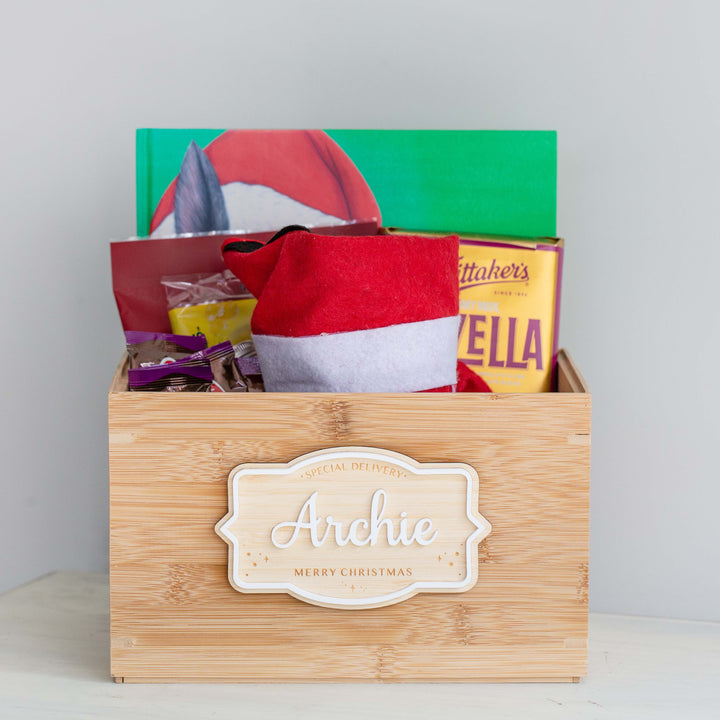 The Personalised Christmas Eve Box is a festive keepsake, featuring a Santa hat, green book, yellow Whitaker’s Hazella chocolate box, and snacks—all beautifully arranged against a plain light background.