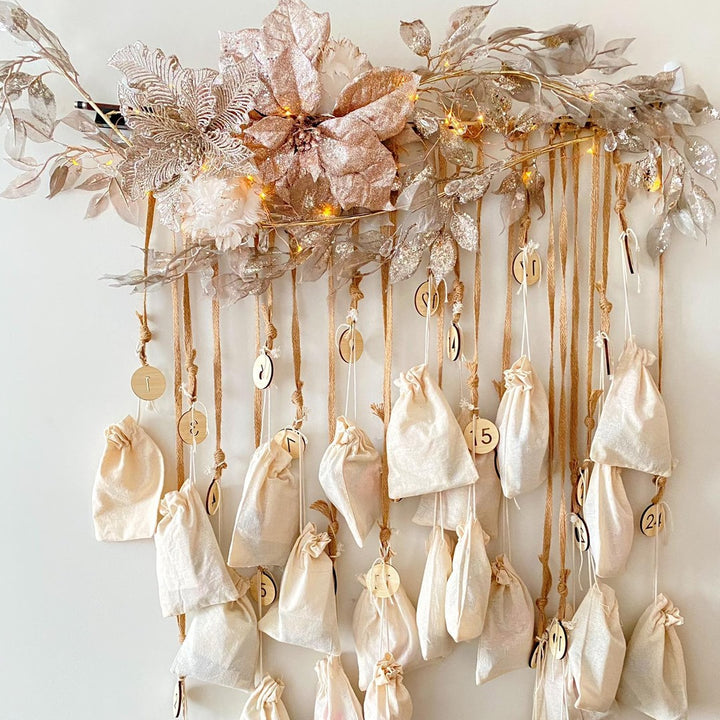The Advent Calendar Tags Set with numbered cream fabric pouches hanging from gold ribbons, decorated with champagne leaves, flowers, and warm fairy lights against a white wall.