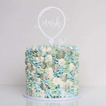 A round cake with blue, green, and white floral icing is topped with a Balloon Personalised Cake Topper (other colours available) displaying "mash" in cursive, set against a plain white background.