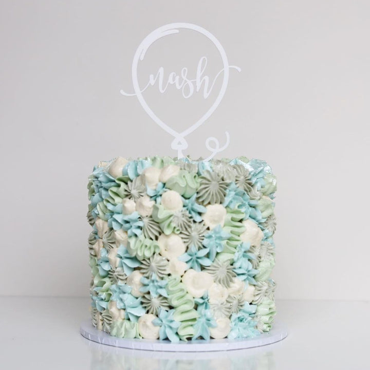 A round cake with blue, green, and white floral icing is topped with a Balloon Personalised Cake Topper (other colours available) displaying "mash" in cursive, set against a plain white background.