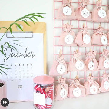 A December calendar page displayed beside a pink bamboo advent calendar with fabric pouches, the Advent Calendar Tags Set, and a jar of red and white items, arranged on a white surface.