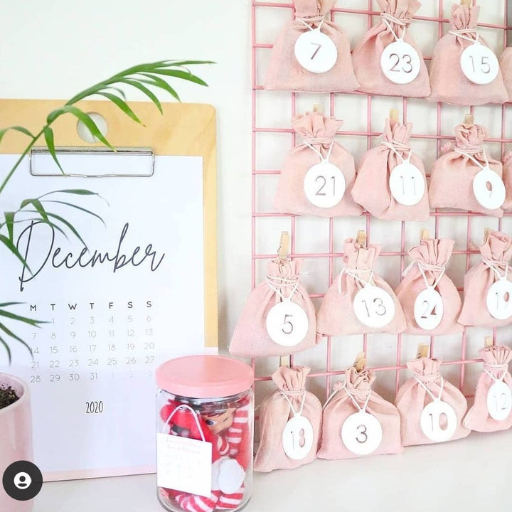 A December calendar page displayed beside a pink bamboo advent calendar with fabric pouches, the Advent Calendar Tags Set, and a jar of red and white items, arranged on a white surface.
