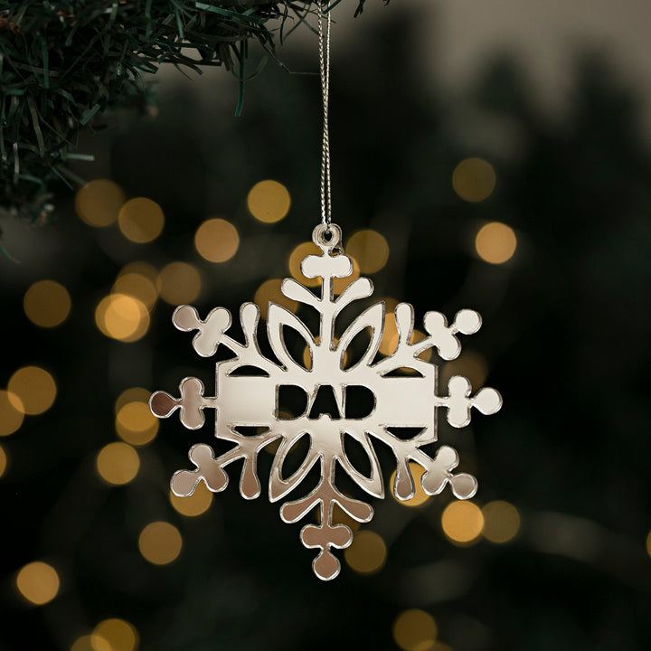 A Personalised Snow Stars ornament with "DAD" in the center hangs from a tree branch, highlighting unique New Zealand made Christmas ornaments with softly glowing lights in the background.