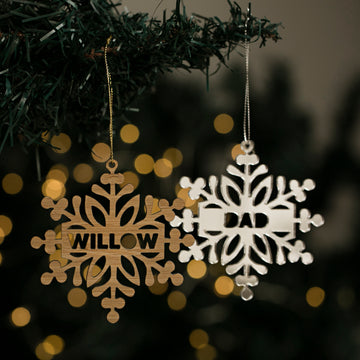 Two Personalised Snow Stars Christmas ornaments hang on a branch—one wooden with "WILLOW," another white with "DAD." Warm holiday lights blur in the background, highlighting these New Zealand made decorations.