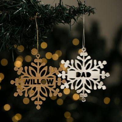 Two Personalised Snow Stars Christmas ornaments hang on a branch, one wooden with "WILLOW," another white with "DAD." Warm holiday lights blur in the background. New Zealand made.