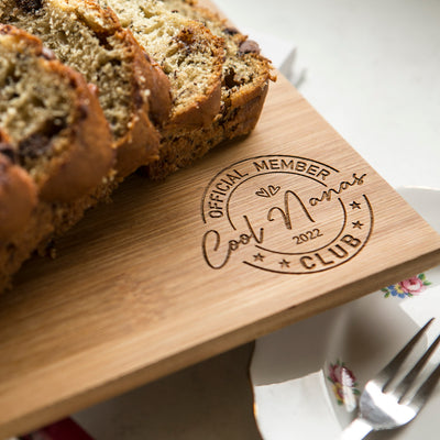 A sliced loaf of bread sits on a Personalised Bamboo Platter/Serve Board engraved with "Official Member Cool Nanas Club 2022." A floral plate and fork peek in, making it an ideal New Zealand gift for special moments. Two board sizes available.