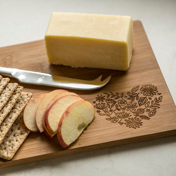 A block of cheese, whole-grain crackers, and apple slices are served on a personalised bamboo platter with a floral engraving. A cheese knife sits beside the cheese—an ideal gift that New Zealand food lovers will cherish. Two sizes available.