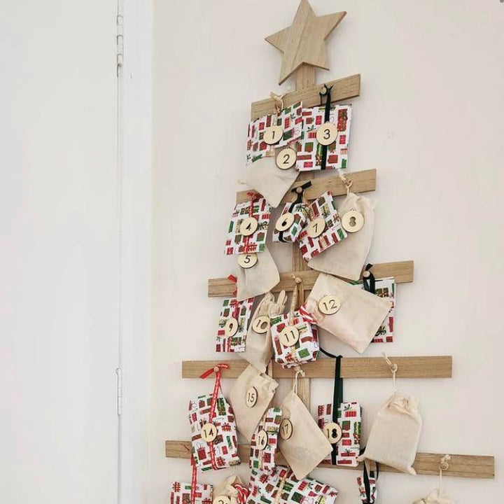 A wooden Christmas tree-shaped frame on a wall decorated with the Advent Calendar Tags Set, including numbered gift bags and small presents, topped with a wooden star.
