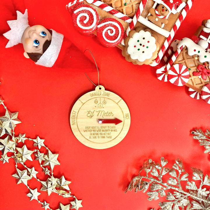 A flat lay of Christmas decor on a red background, featuring the Elf Meter Christmas Bauble, a toy elf head, gingerbread house decorations, gold stars, and glittery silver foliage.