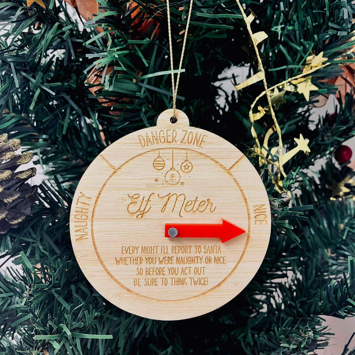 The Elf Meter Christmas Bauble hangs on a tree, its red arrow pointing to “Nice.” The custom bauble features sections like “Naughty,” “Danger Zone,” and “Nice,” amid pine needles and pine cones for a festive touch.