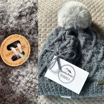 Close-up of a Removable PomPom Button labelled "Handmade" on chunky grey knit fabric, beside a grey cable-knit beanie with faux fur pom-pom, finished with a "Sanity Knits WANAKA" tag.