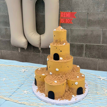 A four-tier sandcastle birthday cake with edible sand, white seashells, and black doorways is topped with a red Flag Personalised Cake Topper that says “Remi is 10.” Large beige balloons decorate the background.