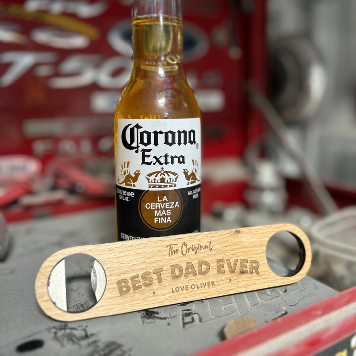 A Personalised Bottle Opener (3 designs available) engraved with The Original BEST DAD EVER LOVE OLIVER rests in front of a Corona Extra beer bottle on a red toolbox, making a practical gift for men. Blurred tools and auto parts appear in the background.