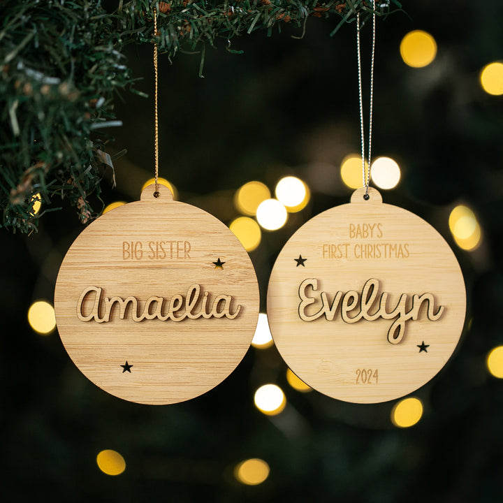 Two Celebration Christmas Baubles hang on a tree—one reads Big Sister Amaelia and the other Baby&