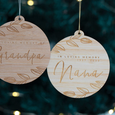 Two wooden ornaments with engraved text 'Grandpa' and 'Mama' against a Christmas tree background.