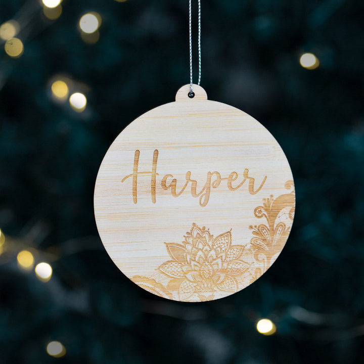 The Lace Christmas Bauble is a round, eco-friendly bamboo ornament featuring the name “Harper” and floral engravings, hanging on a string with blurred holiday lights in the background.