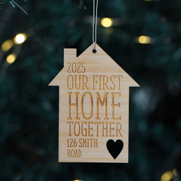 The First Home Christmas Bauble, a wooden house-shaped ornament with "2025 Our First Home Together 126 Smith Road" and a heart engraved, hangs on a string—making an ideal housewarming gift against blurred festive lights.