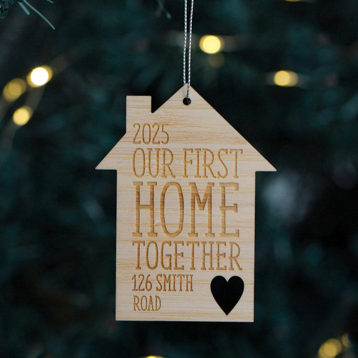 The First Home Christmas Bauble, a wooden house-shaped ornament with "2025 Our First Home Together 126 Smith Road" and a heart engraved, hangs on a string—making an ideal housewarming gift against blurred festive lights.