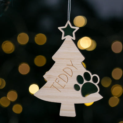 Wooden Christmas tree ornament with 'TEDDY' and a paw print, hanging against a blurred light background.