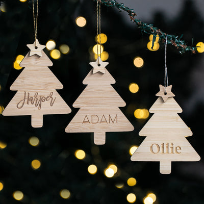 Three wooden Christmas tree ornaments with names 'Harper', 'Adam', and 'Ollie' against a blurred festive background.