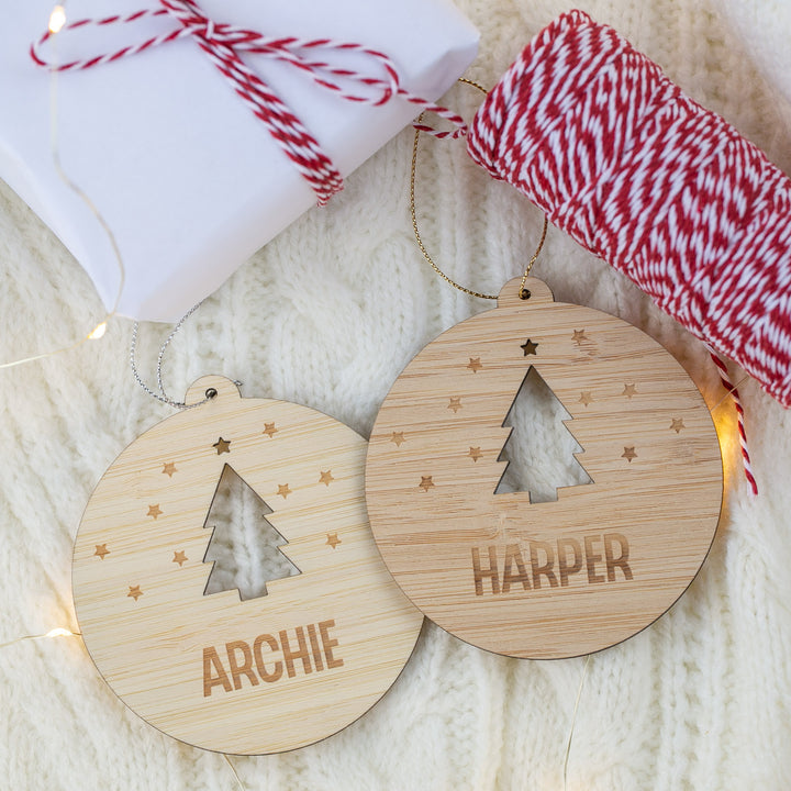 Two wooden Christmas ornaments with cut-out tree shapes and engraved names on a textured surface.