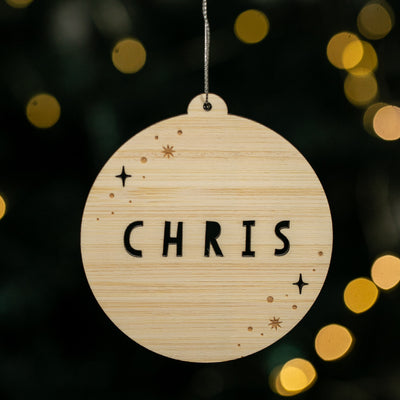 Wooden ornament with 'CHRIS' engraved, hanging against a festive background with lights.