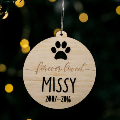 Wooden ornament with paw print and engraved text 'forever loved MISSY 2007-2016' against a blurred Christmas tree background.