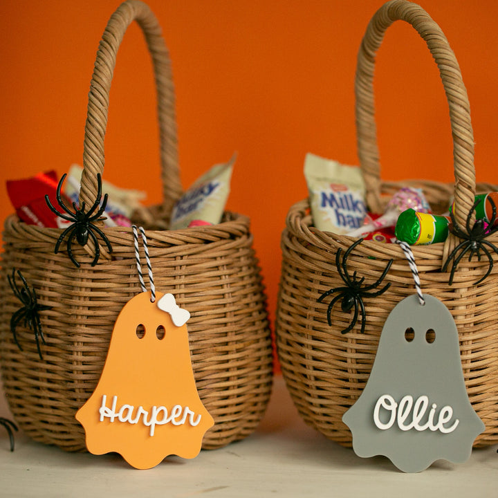 Two wicker baskets filled with Halloween candy, each decorated with black plastic spiders and a personalised acrylic Halloween tag reading Harper and Ollie, set against an orange background.