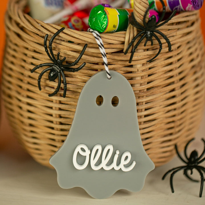 A wicker basket filled with colourful candies and plastic spiders, featuring a Personalised Acrylic Halloween Tag in grey, customised with the name Ollie in white, hanging from the handle.