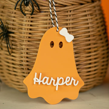 A Personalised Acrylic Halloween Tag in orange ghost shape, customised with "Harper" in white lettering and a white bow, hangs before a wicker basket decorated with black plastic spiders.